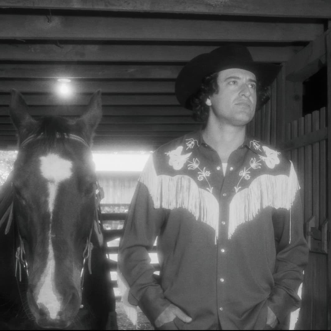 Joey Myron in a black hat and fringe shirt beside a horse in a barn
